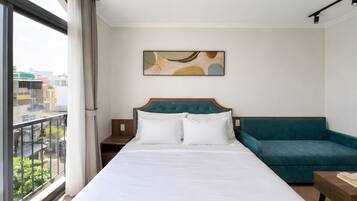 A hotel room with a large bed, a teal sofa, and a city view through a window.