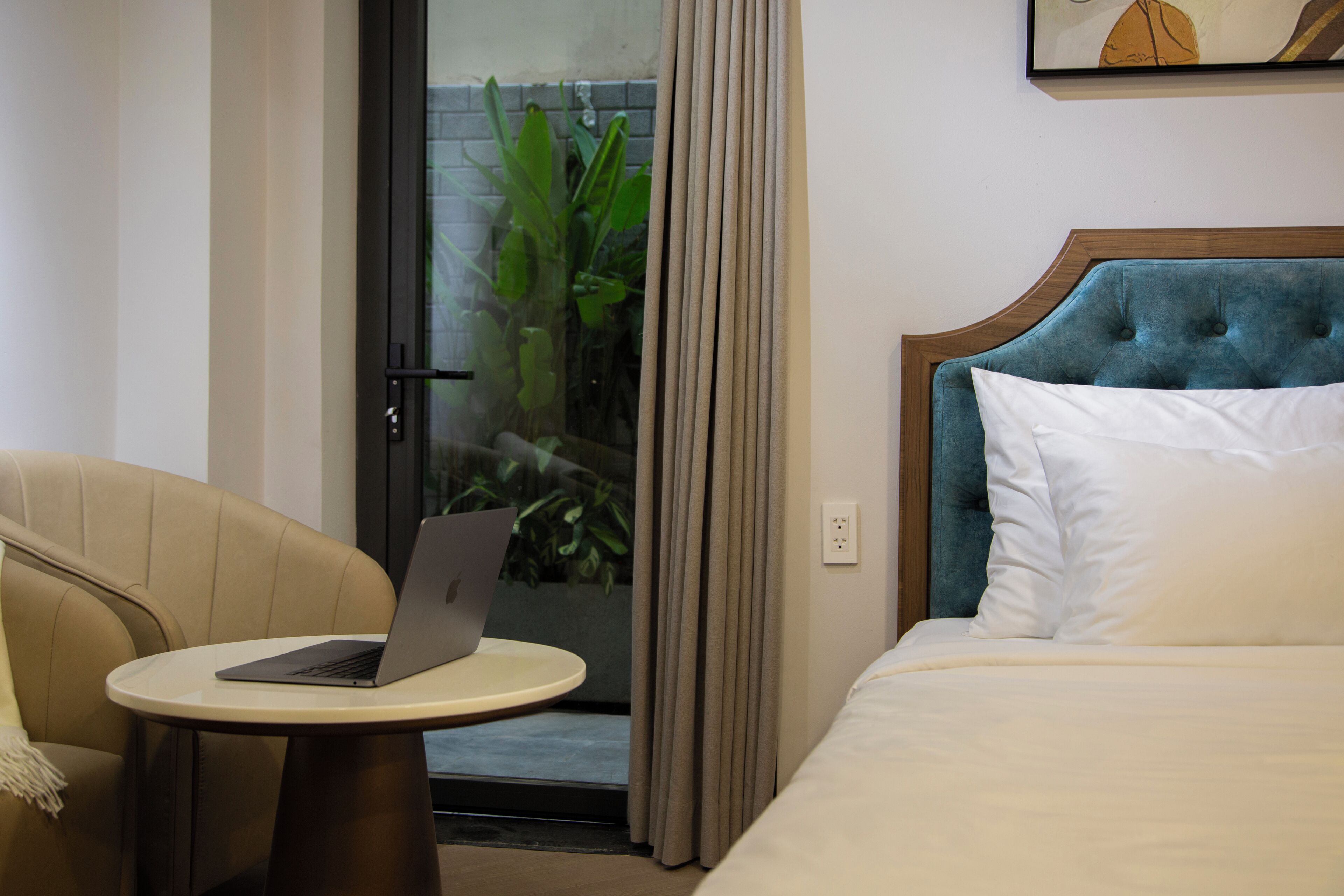 A hotel room with a bed, a chair, a small table with a laptop, and a view of greenery through a glass door.