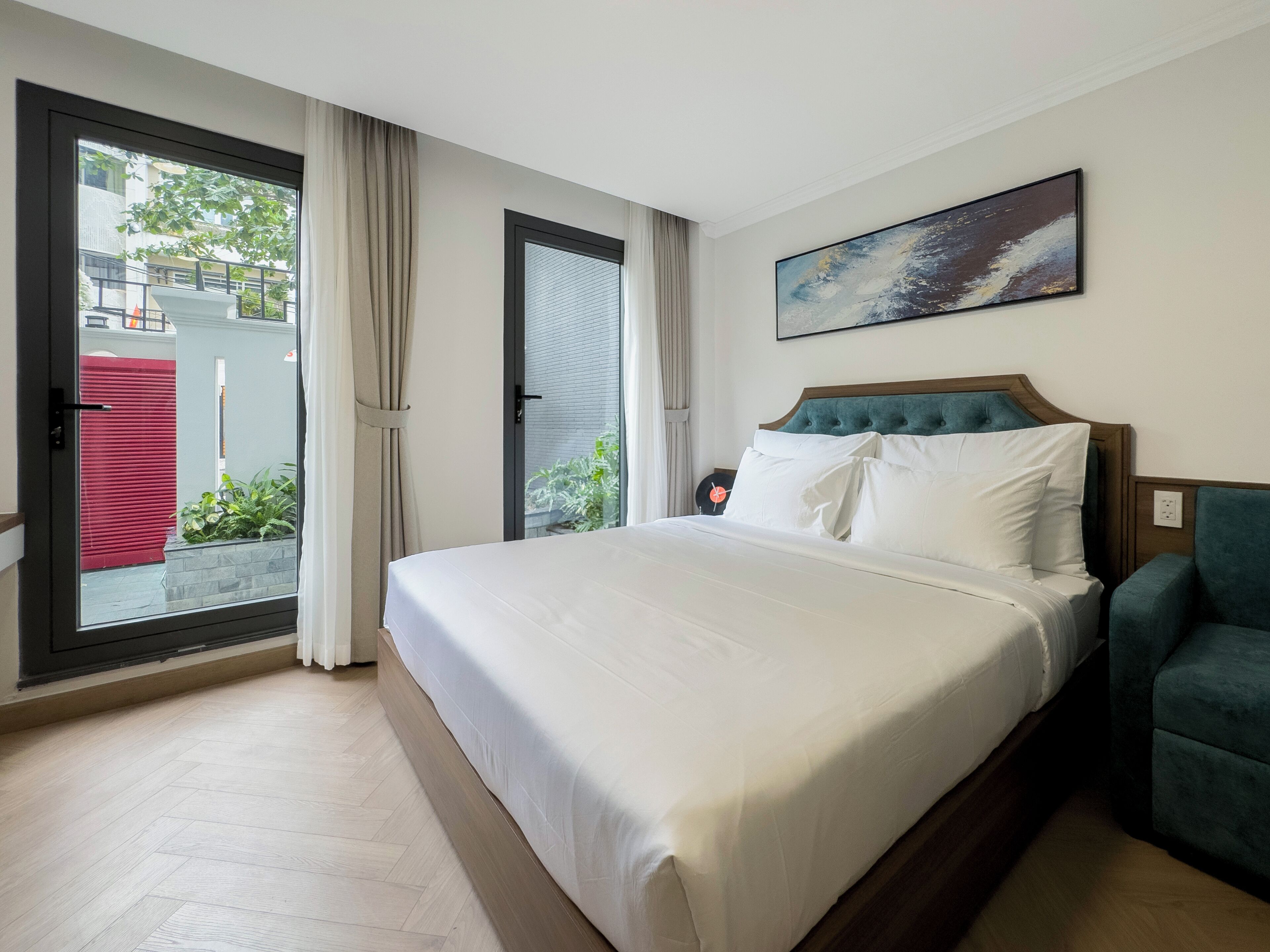 A hotel room with a large bed, a view through a sliding door, a painting on the wall, and a teal armchair.