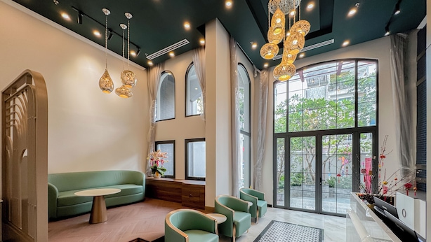 Un intérieur moderne avec un canapé vert, une petite table et un coin salon avec des chaises. De grandes fenêtres sont ornées de rideaux, le sol est recouvert d’un motif décoratif et un luminaire orne le plafond.