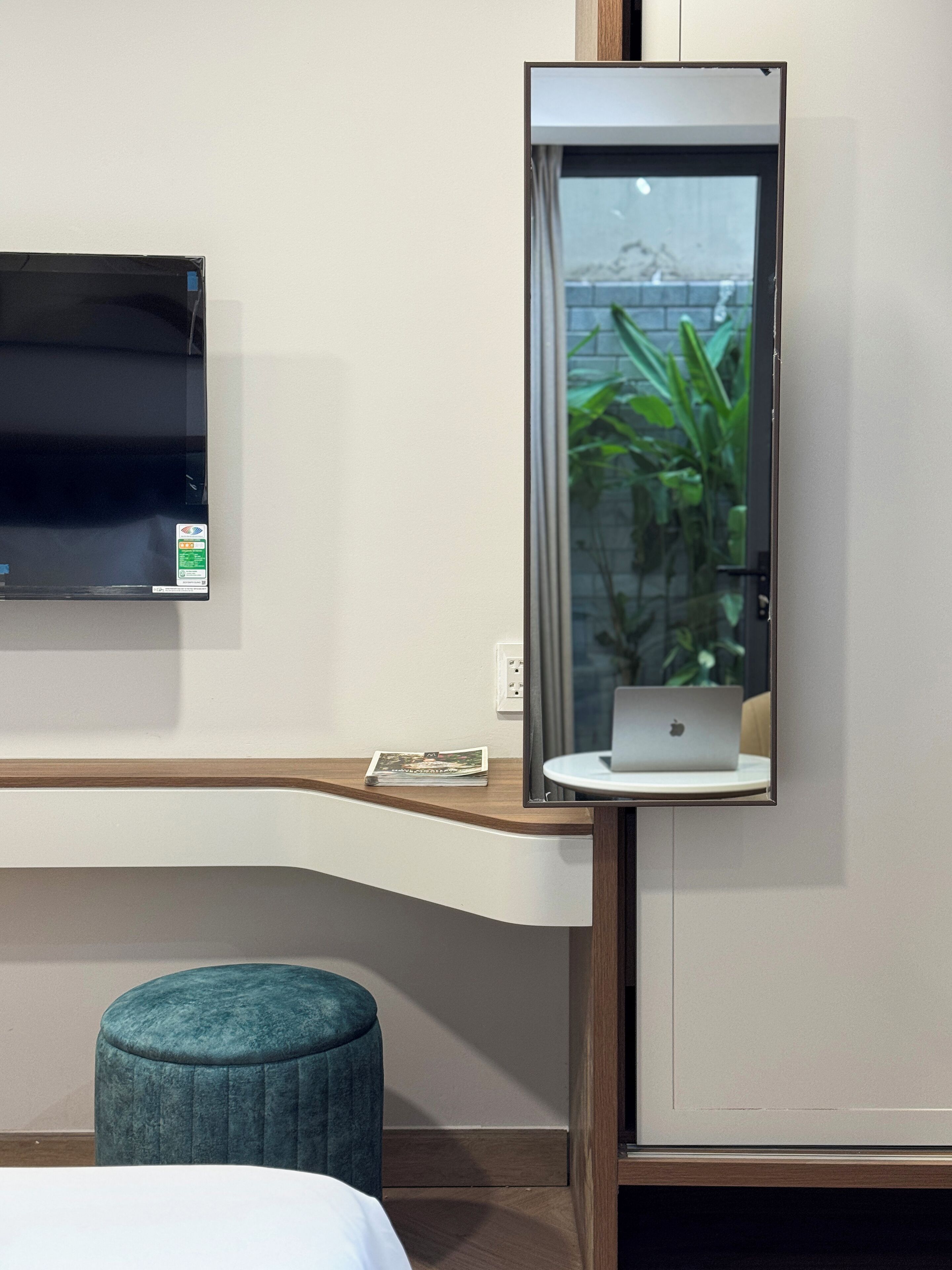 Ein modernes Hotelzimmer mit einem wandmontierten Flachbildfernseher, einem Holztisch mit einem Laptop, einem Türkis-Pouff und einer kleinen Pflanze, die sich im Spiegelbild zeigt.