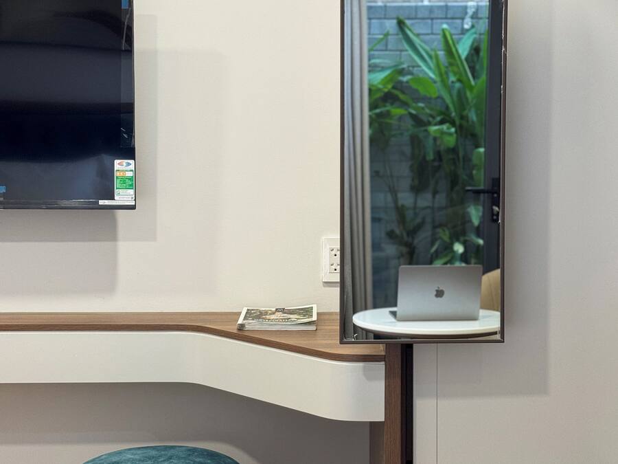 Una habitación de hotel moderna con un televisor de pantalla plana montado en la pared, un escritorio de madera con un ordenador portátil, un reposapiés color turquesa y una planta pequeña visible en el reflejo.