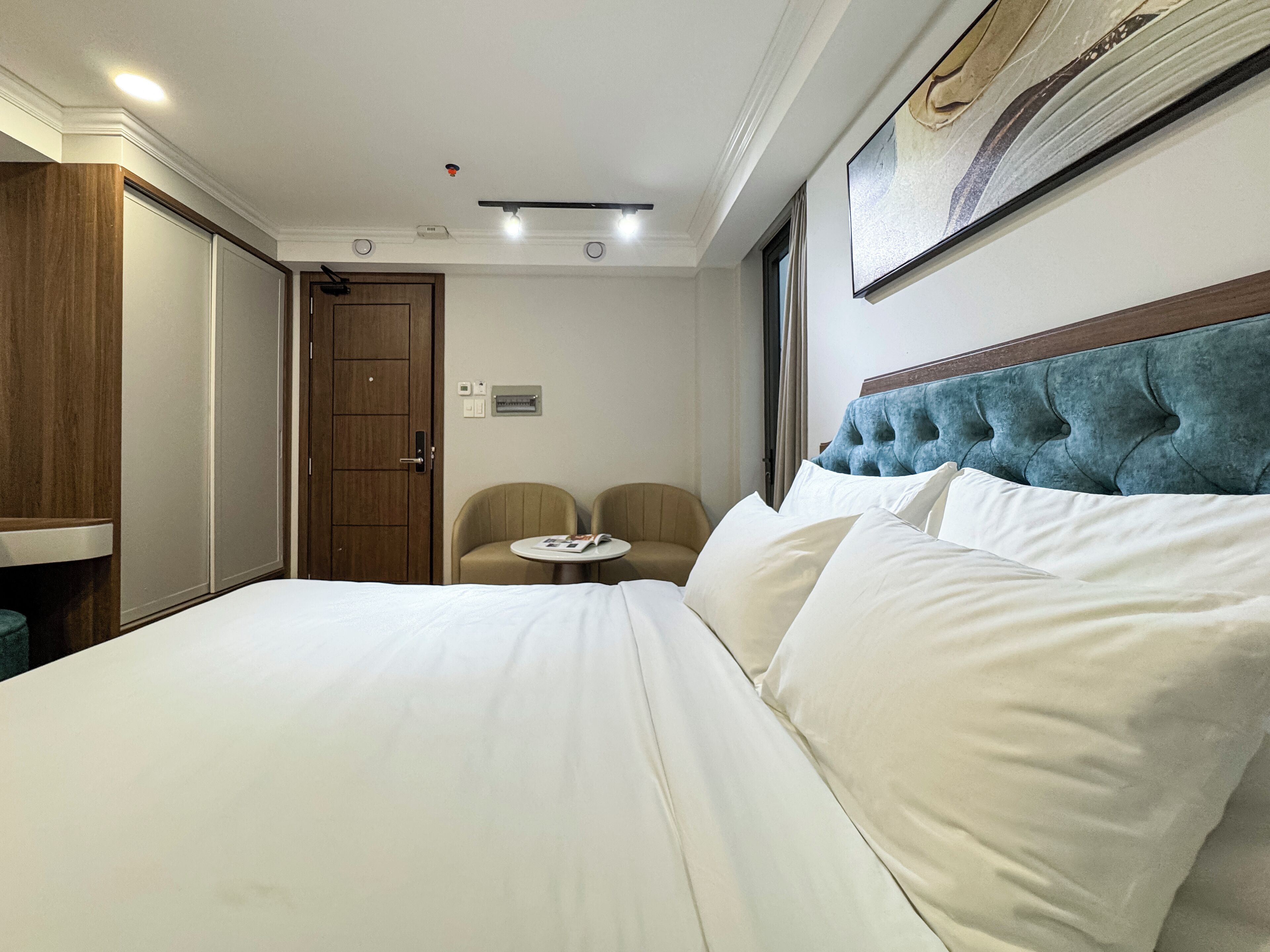 A hotel room with a large bed, a small round table, two chairs, a wooden wardrobe, and a large window.