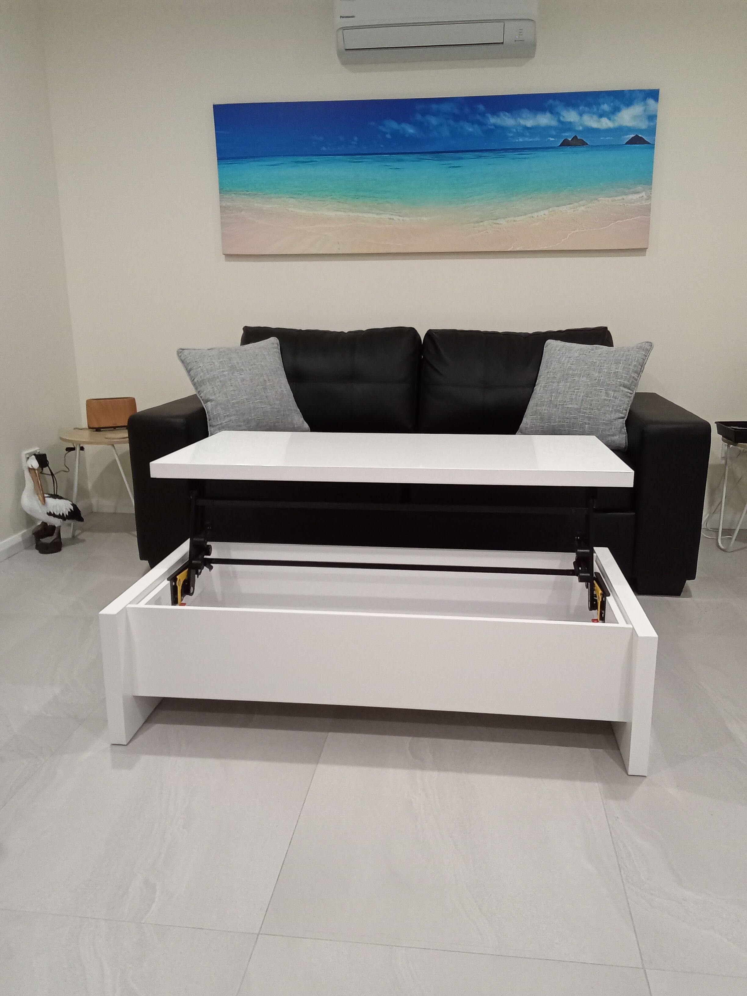 A modern living room with a black sofa, white coffee table, and a painting of a beach scene on the wall.