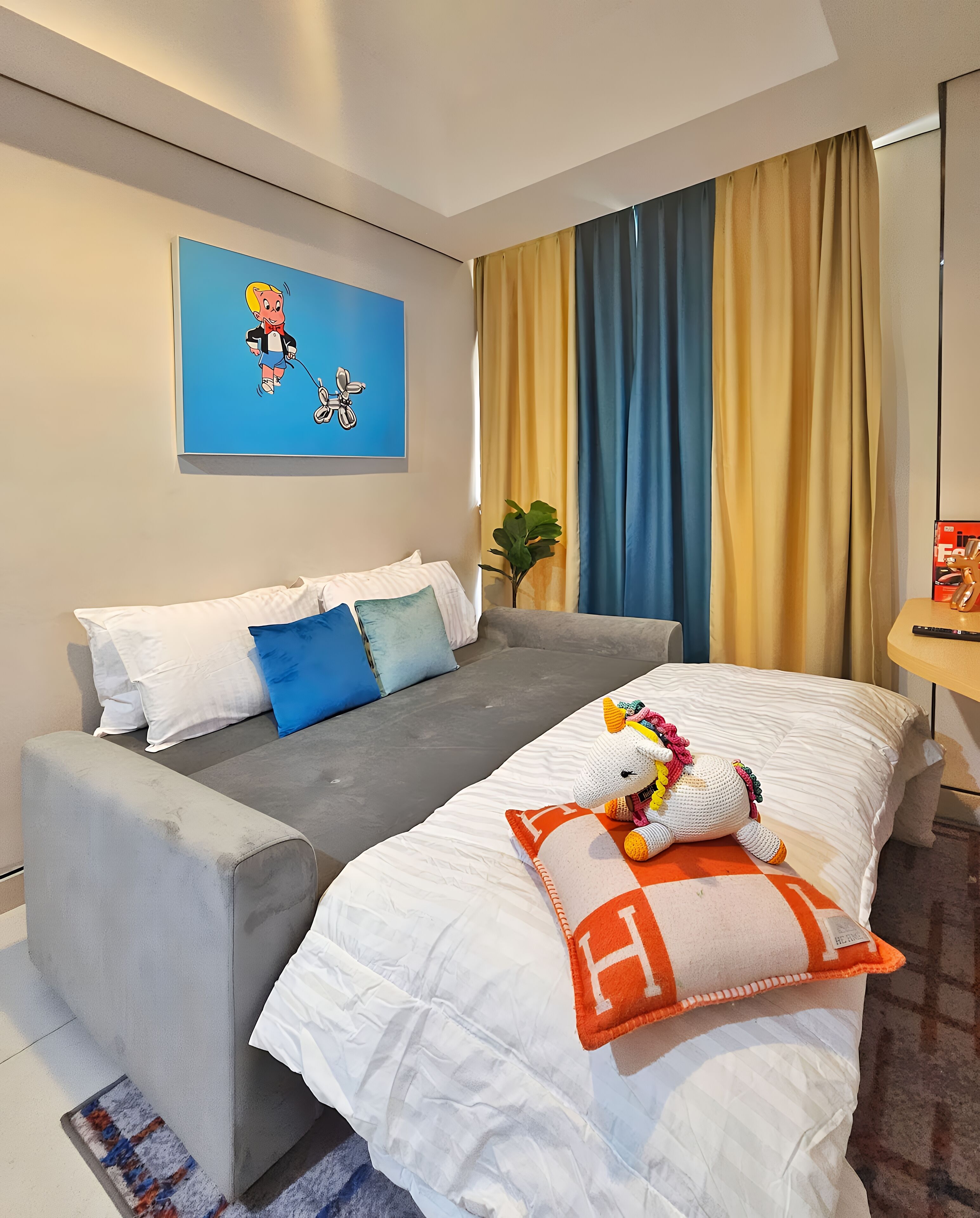 Un cuarto de hotel con un sofá gris, una cama con almohadas blancas y anaranjadas, una obra de arte azul en la pared y una planta cerca de la ventana.