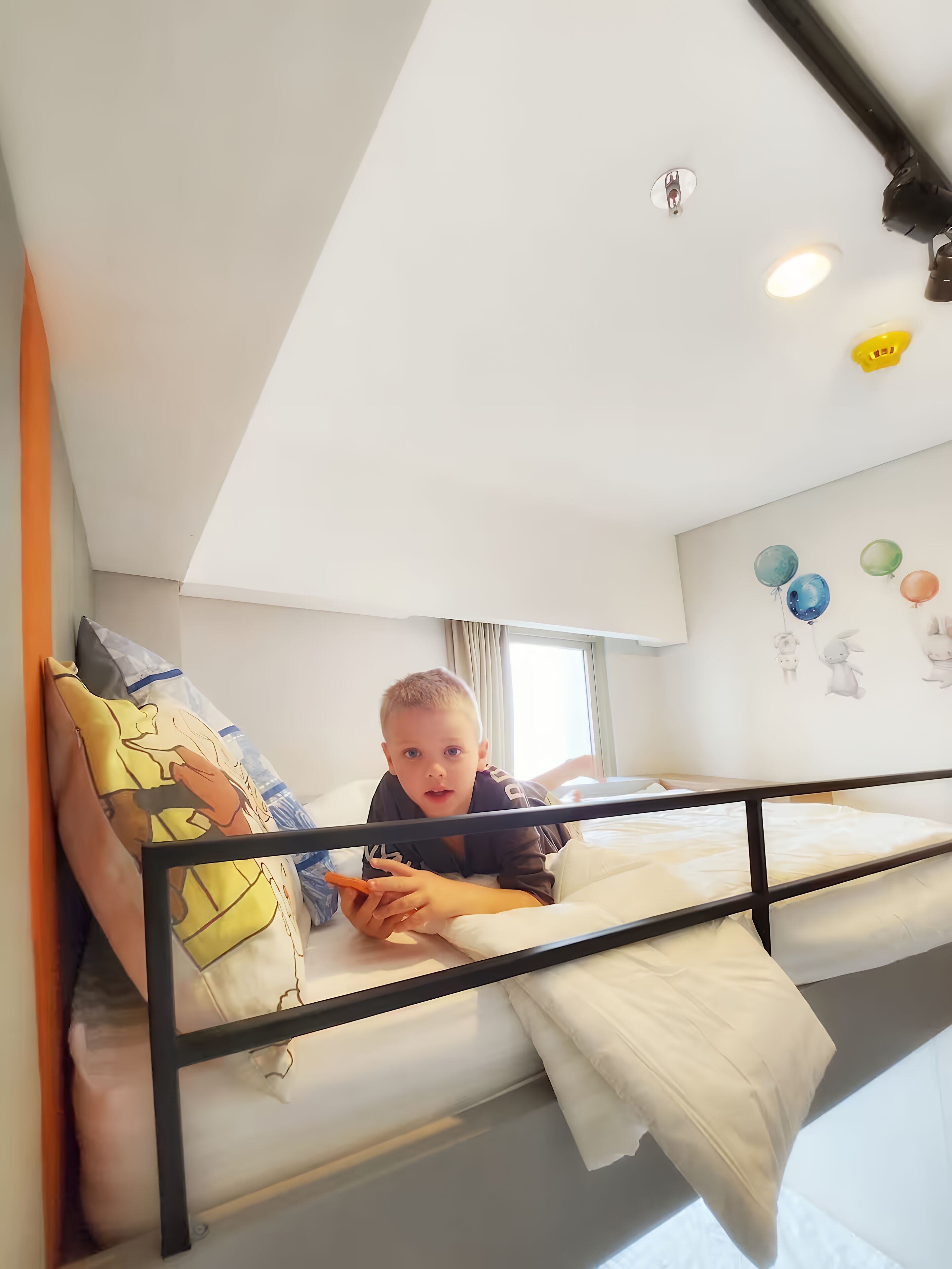 Ein Kind liegt auf einem Etagenbett in einem Zimmer mit weißer Decke, einem Fenster mit Vorhängen und Wanddekorationen, darunter Ballons und Zeichentrickfiguren.