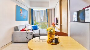A modern living room with a grey sofa, a round table with a vase of flowers, and a large window with a city view. - Hava By SanLiving - 1 BR - Taman Anggrek (Jakarta)