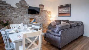 A cozy living room with a stone wall, a grey sofa, a dining table set for four, and a flat-screen TV mounted on the wall. - Tide & Tee - 2 Bedroom Apartment - Penally - Tenby (Tenby)