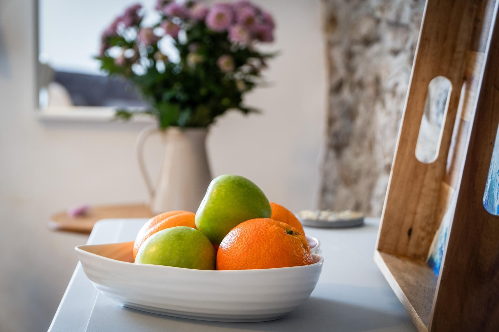 A bowl of fruit on a table with a vase of flowers in the background.