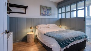 A bedroom with a bed, two bedside lamps, a round side table, a painting on the wall, and a window with blinds. - Tide & Tee - 2 Bedroom Apartment - Penally - Tenby (Tenby)