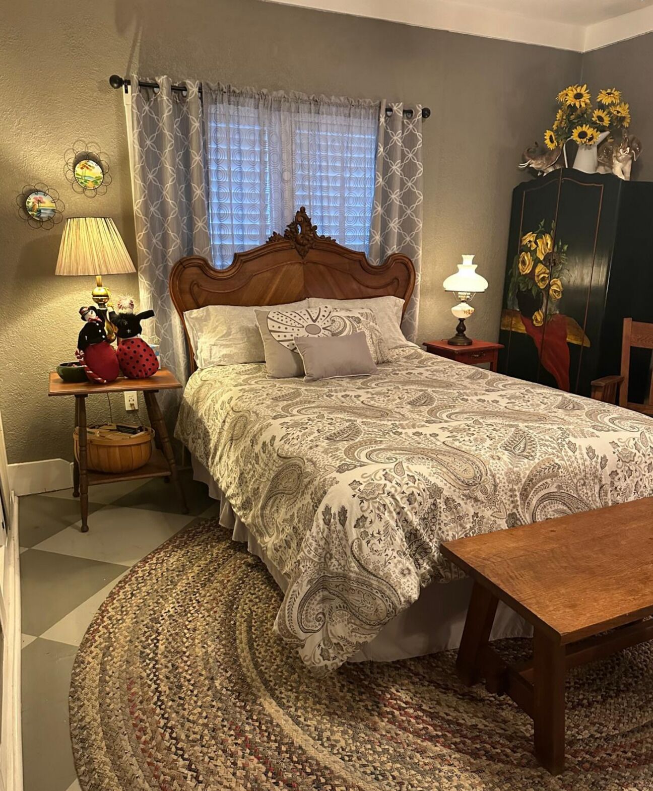 Un dormitorio con una cama grande, una mesita de noche de madera, una lámpara, una pequeña mesa con figuritas, un aparador decorativo y una alfombra estampada.