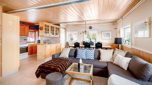 Un ampio soggiorno con soffitto in legno, un divano, un tavolino in vetro e una cucina sullo sfondo.
