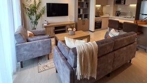 A modern living room with a grey sofa, a flat-screen TV, and a wooden coffee table. There is a kitchen area with a microwave and a dining table with chairs. - 1 Bedroom Furnished to let in GTC Westlands. (Nairobi)