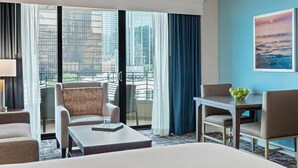 Een hotelkamer met een groot bed, twee fauteuils, een kleine tafel en uitzicht op een stadsgezicht door grote ramen.