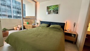 A bedroom with a large bed, a nightstand, a bedside table, a window with a view of buildings, and a potted plant. - Luxurious, modern, amongst it all (South Yarra)