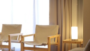 A hotel room with two wooden chairs, a bedside table with a lamp, and a large window with curtains.