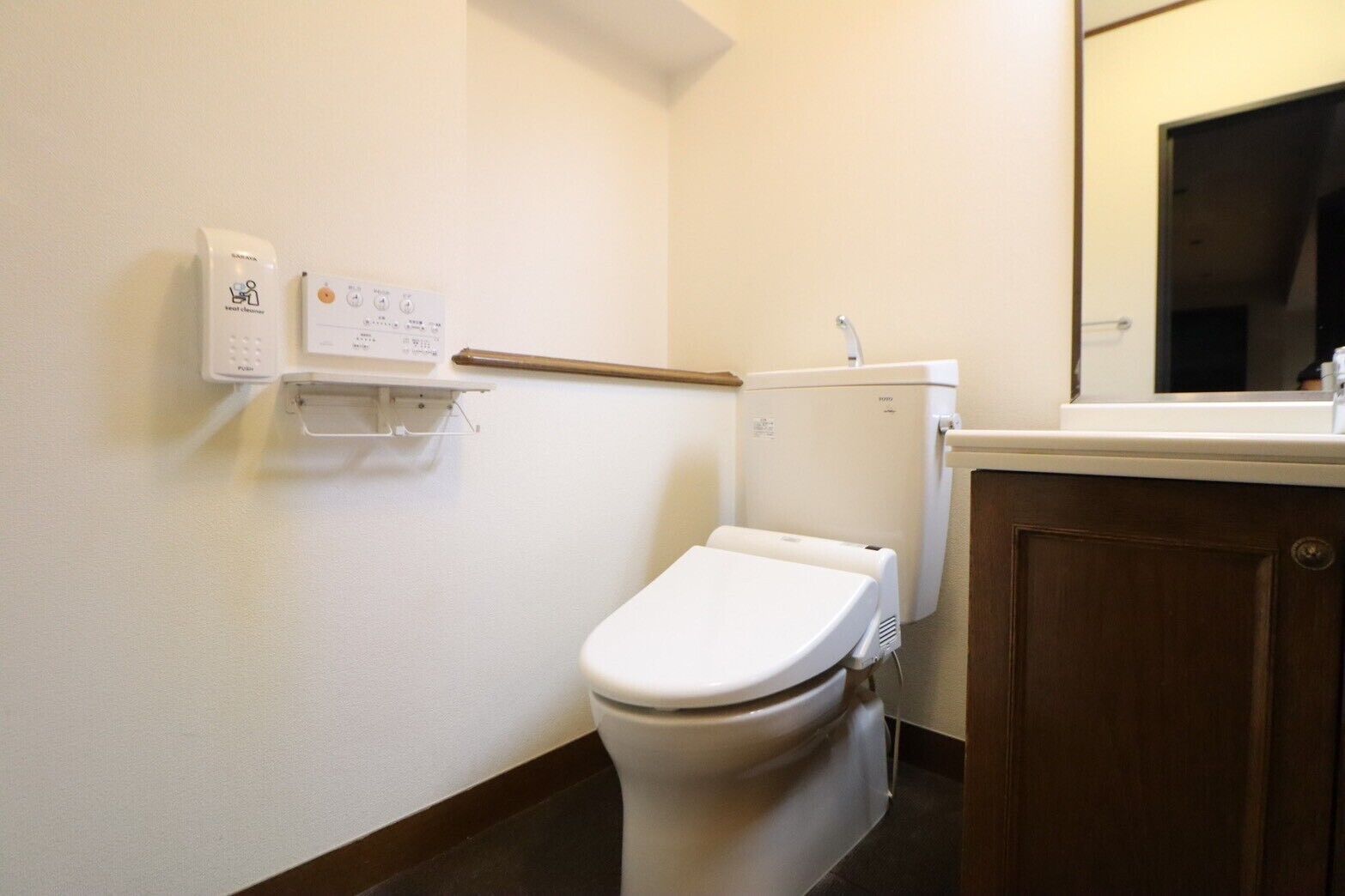 Une petite salle de bain propre, équipée de toilettes, d’un sèche-mains et d’un distributeur de savon fixé au mur.