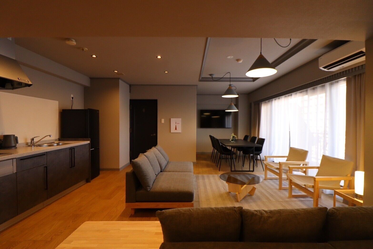 A modern living room with a sofa, a dining table with chairs, a kitchen area with a sink and stove, and a large window with curtains.