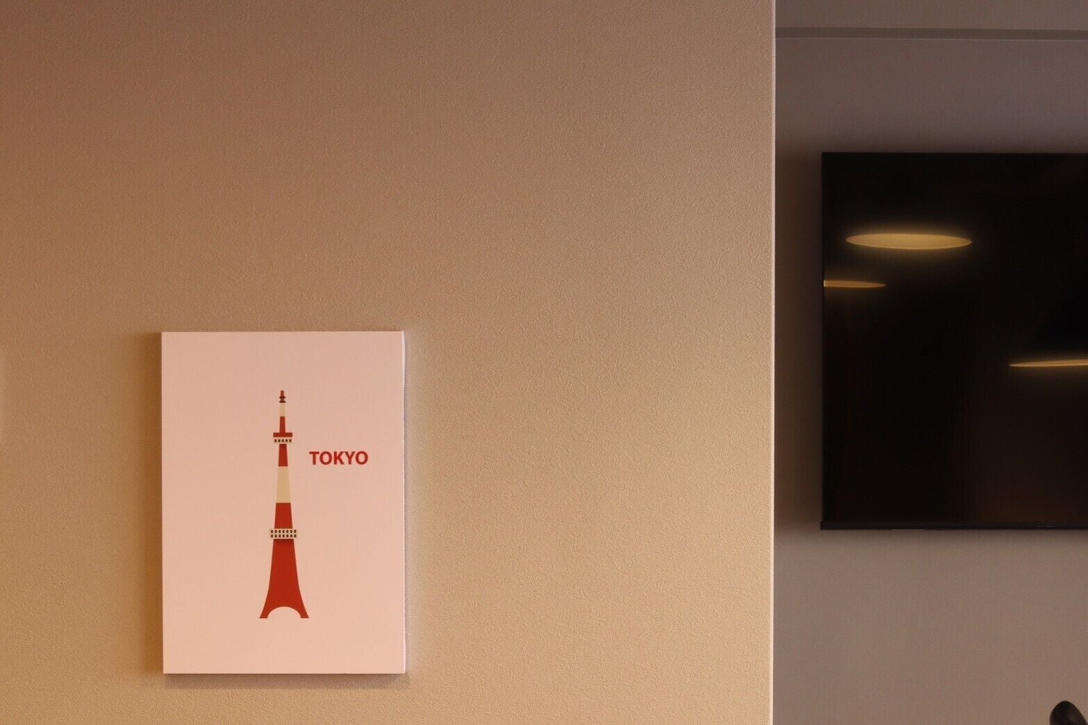 Eine gerahmte Illustration eines Tokioler Wahrzeichens an einer beigen Wand, nebenan eine Wand mit einem flachbildfernseher.