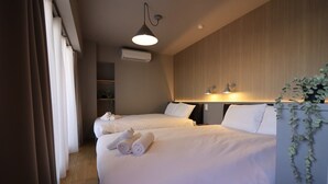 A hotel room with two beds, a bedside table with a lamp, an air conditioning unit, and a plant.