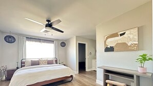 Un dormitorio con cama, escritorio, silla, ventilador de techo, ventana con cortinas y un cuadro en la pared.