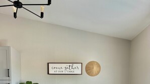 Un área de comedor con una mesa cubierta con un mantel color beige, rodeada de sillas blancas, un jarrón decorativo con flores y un letrero en la pared.
