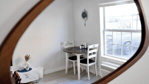 A dining area with a round table, white chairs, a clock on the wall, and a window with a view of parked cars. - Redspeck Luxury Furnished Flats (Aztec)