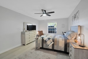 Un dormitorio con cama, ventilador de techo, cómoda, silla, ventana y cuadros en la pared.
