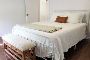 Una cama bien hecha con edredón blanco y almohadas, un banco de madera con cojín y estructura de cama de metal blanco.