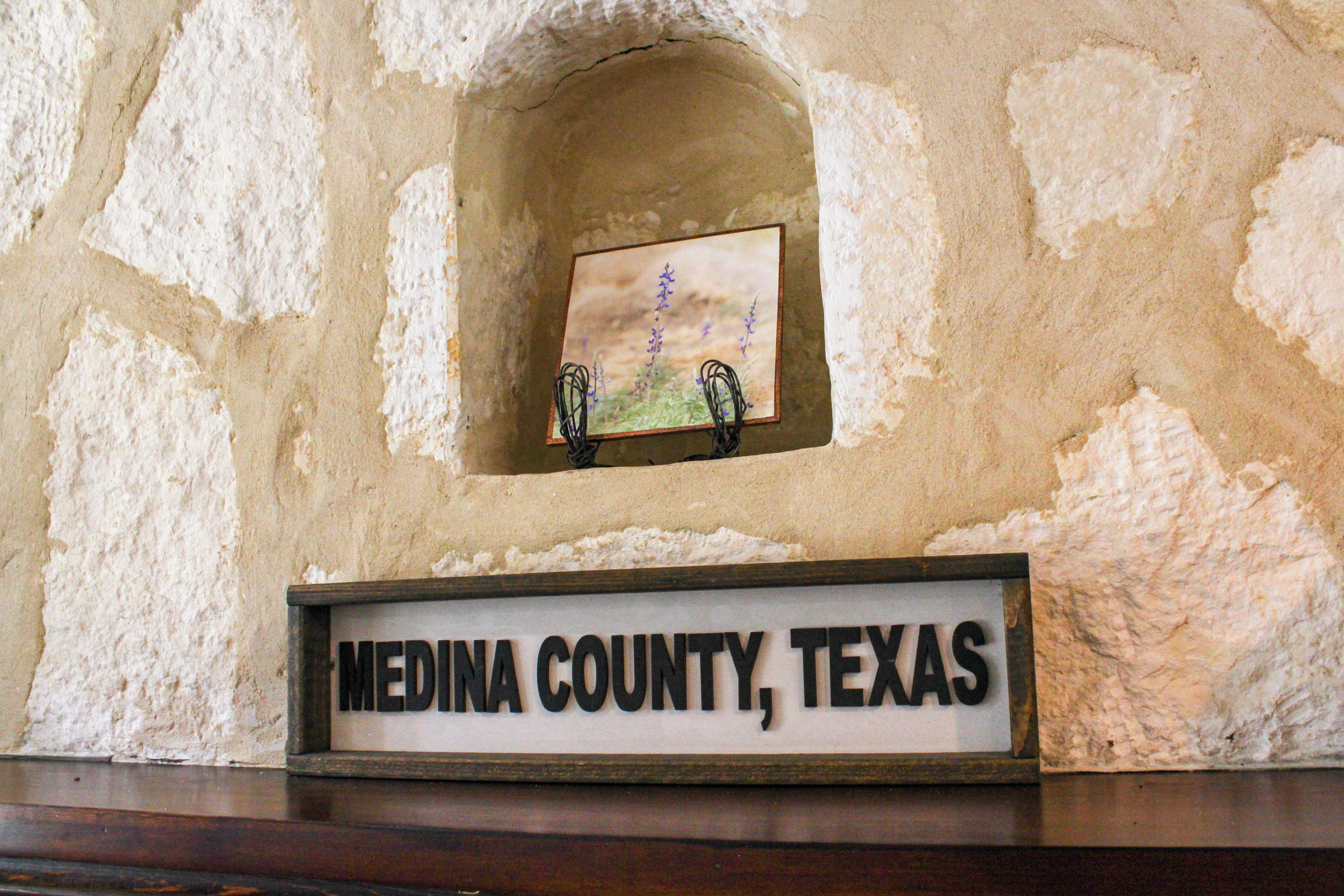 A stone wall with a recessed area containing a framed picture and a sign that reads 'MEDINA COUNTY, TEXAS'.