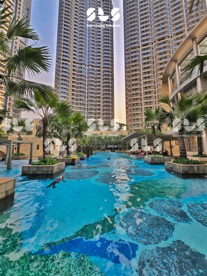 A swimming pool surrounded by tall residential buildings, palm trees, and a clear sky. - Asmara By SanLiving - 1 BR - Direct Mall (Jakarta)