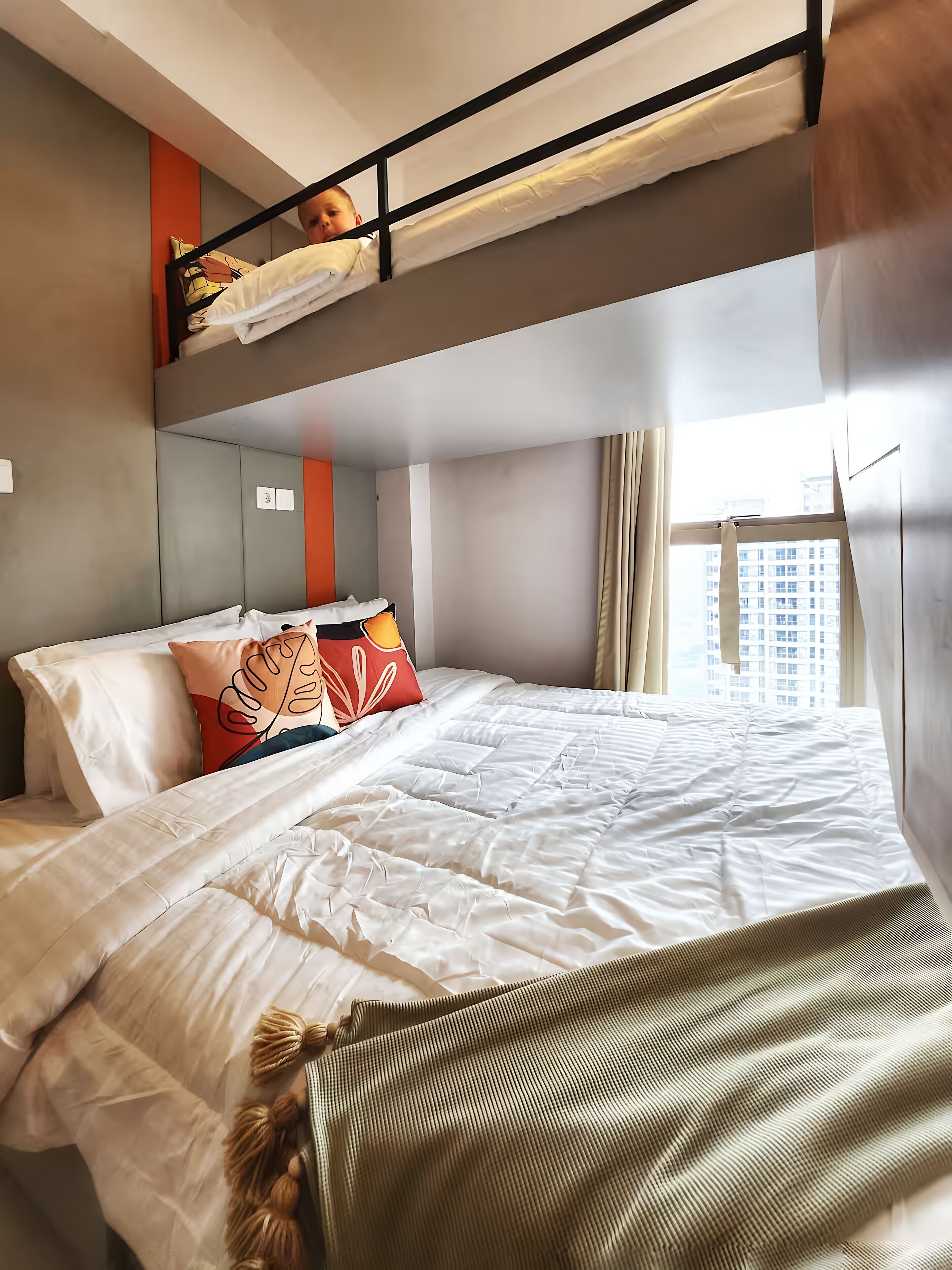 A hotel room with a bunk bed, a large bed, and a window with a city view.