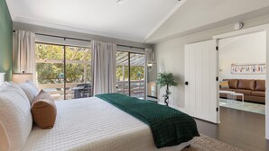 Ein Schlafzimmer mit Bett, Deckenventilator, großem Fenster mit Vorhängen, einer Tür zu einem anderen Raum und Blick ins Freie.