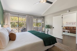 Ein Schlafzimmer mit Bett, Deckenventilator, großem Fenster mit Vorhängen, einer Tür zu einem anderen Raum und Blick ins Freie.