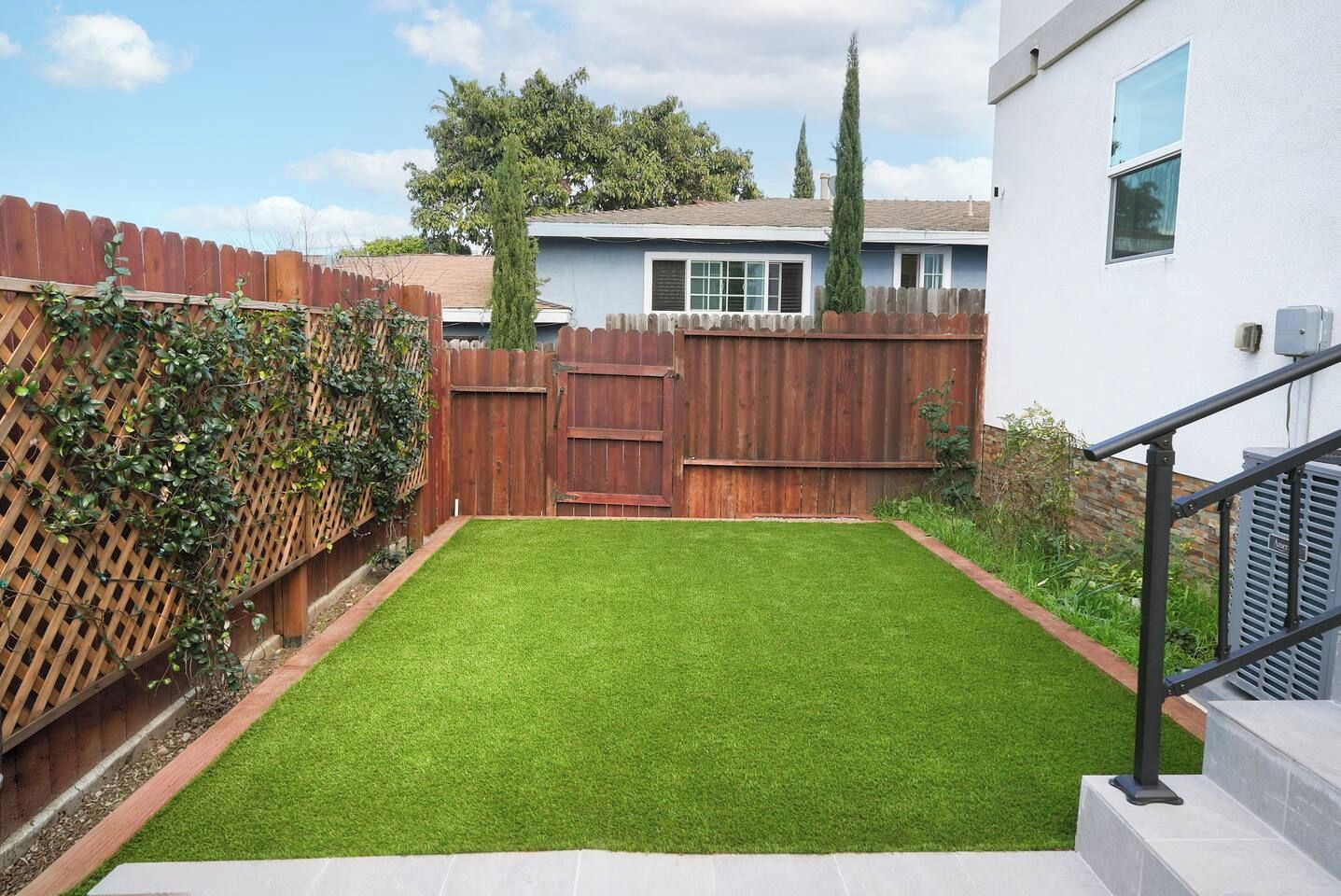 A backyard with a wooden fence, a grassy lawn, and a staircase leading to a white building.