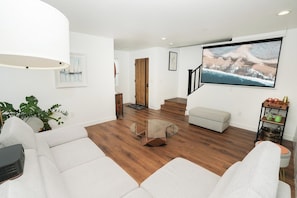 Ein modernes Wohnzimmer mit einem Glastisch, weißen Sofas, Holzboden und einem großen gerahmten Bild an der Wand.