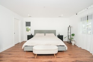 Ein Schlafzimmer mit einem Bett, Nachttischen, einer Bank, einer Stehlampe und einem Fenster mit Vorhängen.