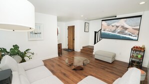 Een moderne woonkamer met een glazen salontafel, witte banken, houten vloeren en een grote tv aan de muur.