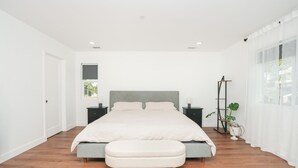 Een moderne slaapkamer met een groot bed, nachtkastjes, een bankje, een vloerlamp en een kamerplant.