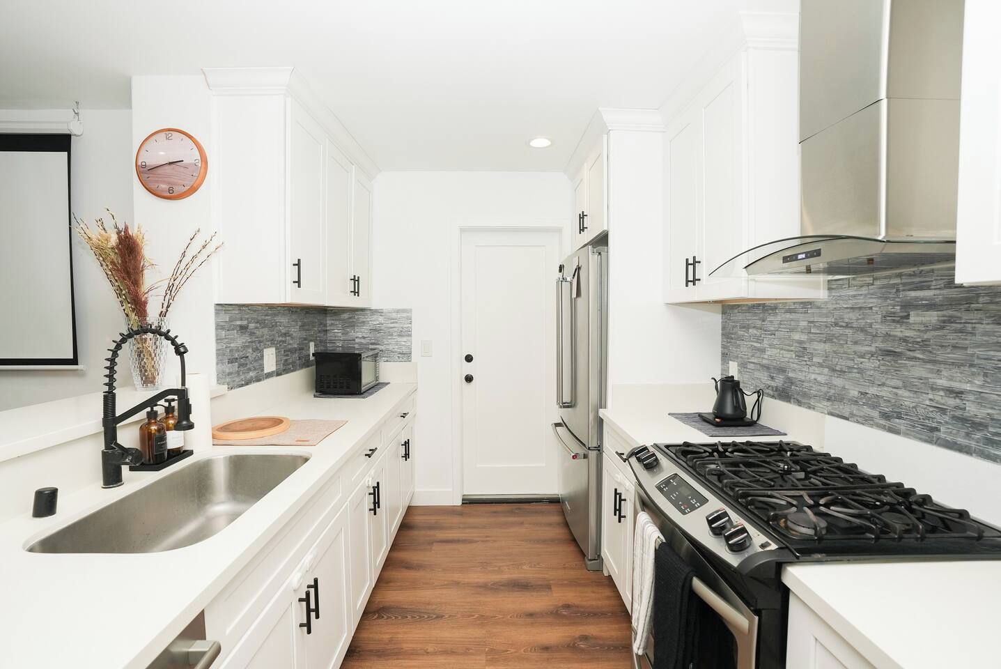 A modern kitchen with white cabinetry, a stainless steel sink, a black faucet, a microwave, a stove, and a clock on the wall.