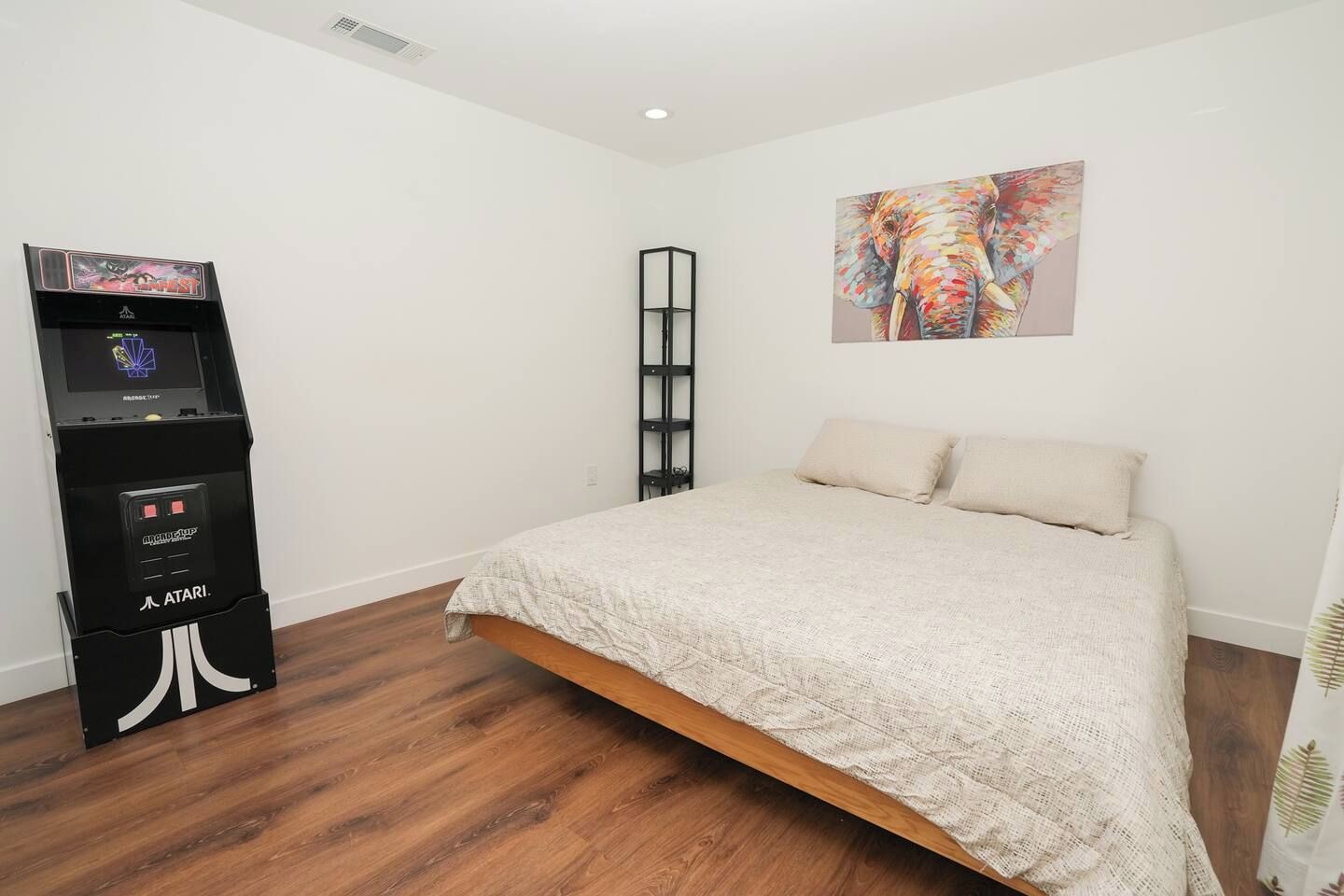 A bedroom with a bed, a gaming console, a bookshelf, and a painting on the wall.