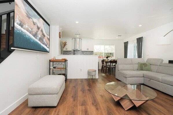 A modern living room with a grey sofa, a glass coffee table, and a dining area with wooden chairs.