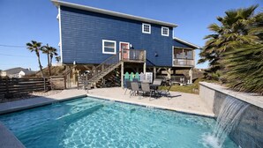 A swimming pool with a waterfall feature, a covered deck with chairs, and a two-story house with a red door. - Private Pool, Large Back Porch, Golf Cart Access (Port Aransas)