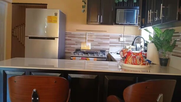 Une cuisine compacte équipée d’un réfrigérateur, d’un four à micro-ondes et d’une cuisinière à gaz. Elle comprend deux tabourets de bar et un évier avec un robinet.