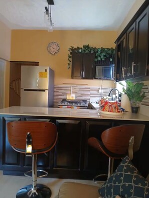 A compact kitchen with a refrigerator, microwave, and gas stove. There are two bar stools and a sink with a faucet. - Susan's Cozy Place (Portmore)