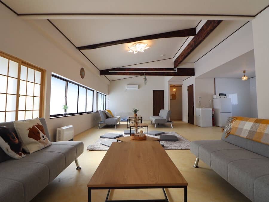 A spacious living room with a wooden coffee table, grey sofas, and large windows providing natural light.