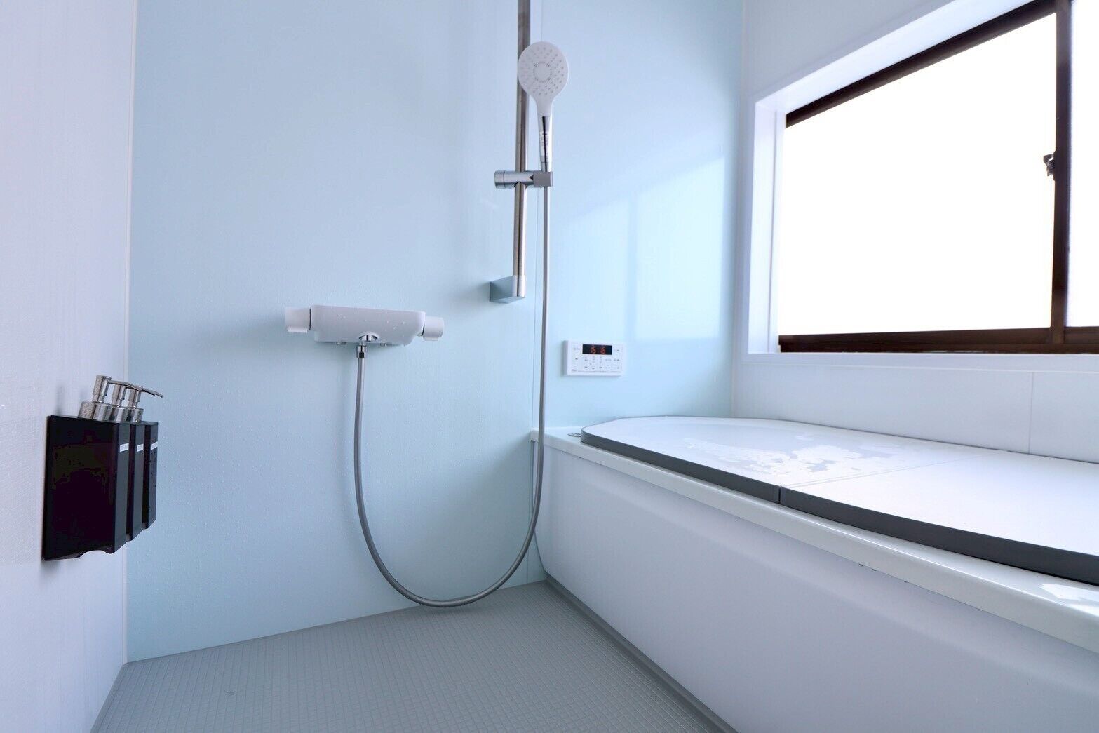 A bathroom with a shower, a soap dispenser, a thermostat, and a window.