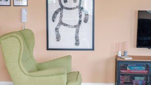 A living room with a green armchair, a blue sofa, a framed abstract artwork, a TV, and a small side table with candles. - Modern Margate Apt 2BR En-suite Parking by Adliv (Margate)