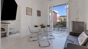 Ein modernes Wohnzimmer mit Esstisch, klaren Stühlen, einer Couch, einem Fernseher und einem Balkon mit Blick auf Gebäude.