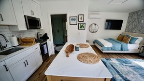 Et moderne køkken med hvide skabe, en mikrobølgeovn og en komfur. Der er et spisebord med en bordskåner og et blåt tæppe. En sofa med puder findes i opholdsstuen.